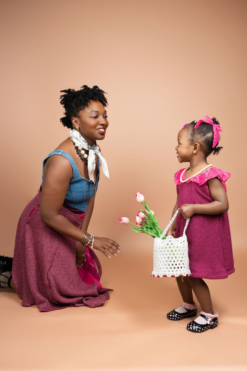 Young girl in pink dress offers tulips to smiling woman kneeling, both in matching burgundy outfits on beige background.