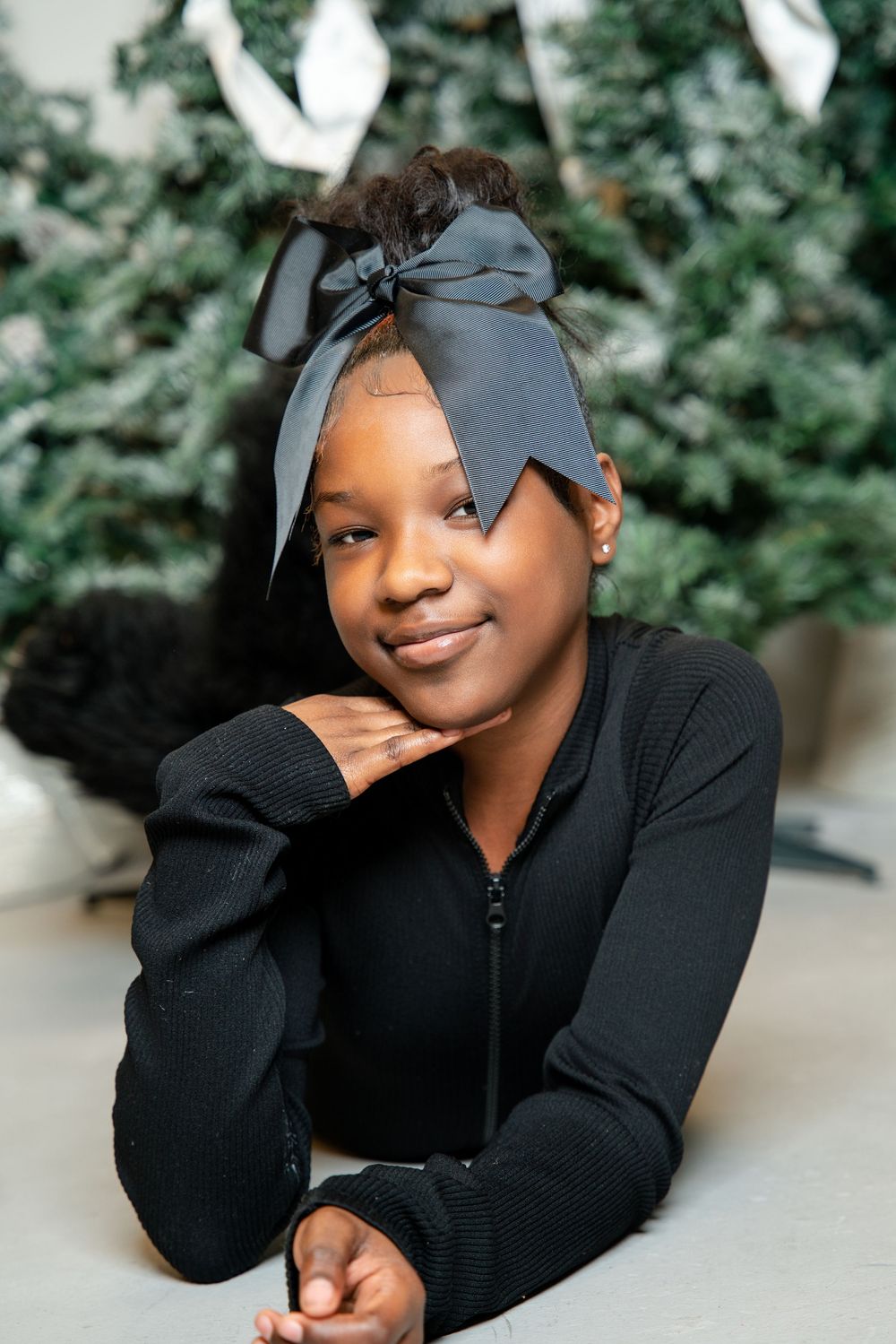 Portrait series in black sweater with gray bow accessory against blurred green foliage background.