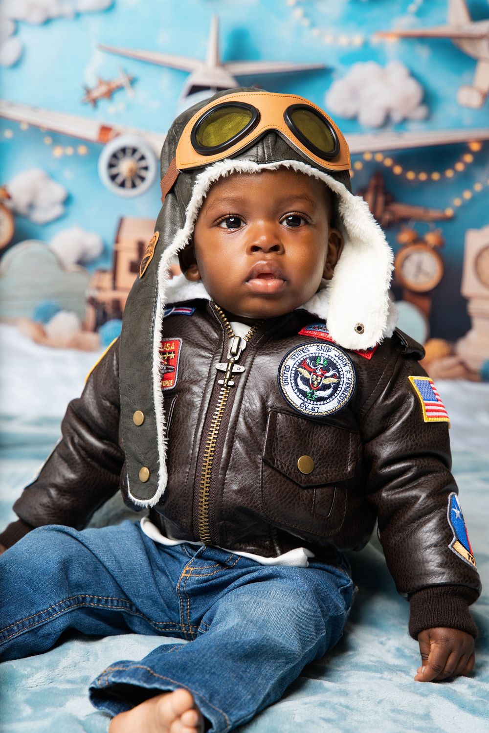 Adorable baby boy in leather pilot jacket with aviator goggles and hat, posed before airplane-themed backdrop.