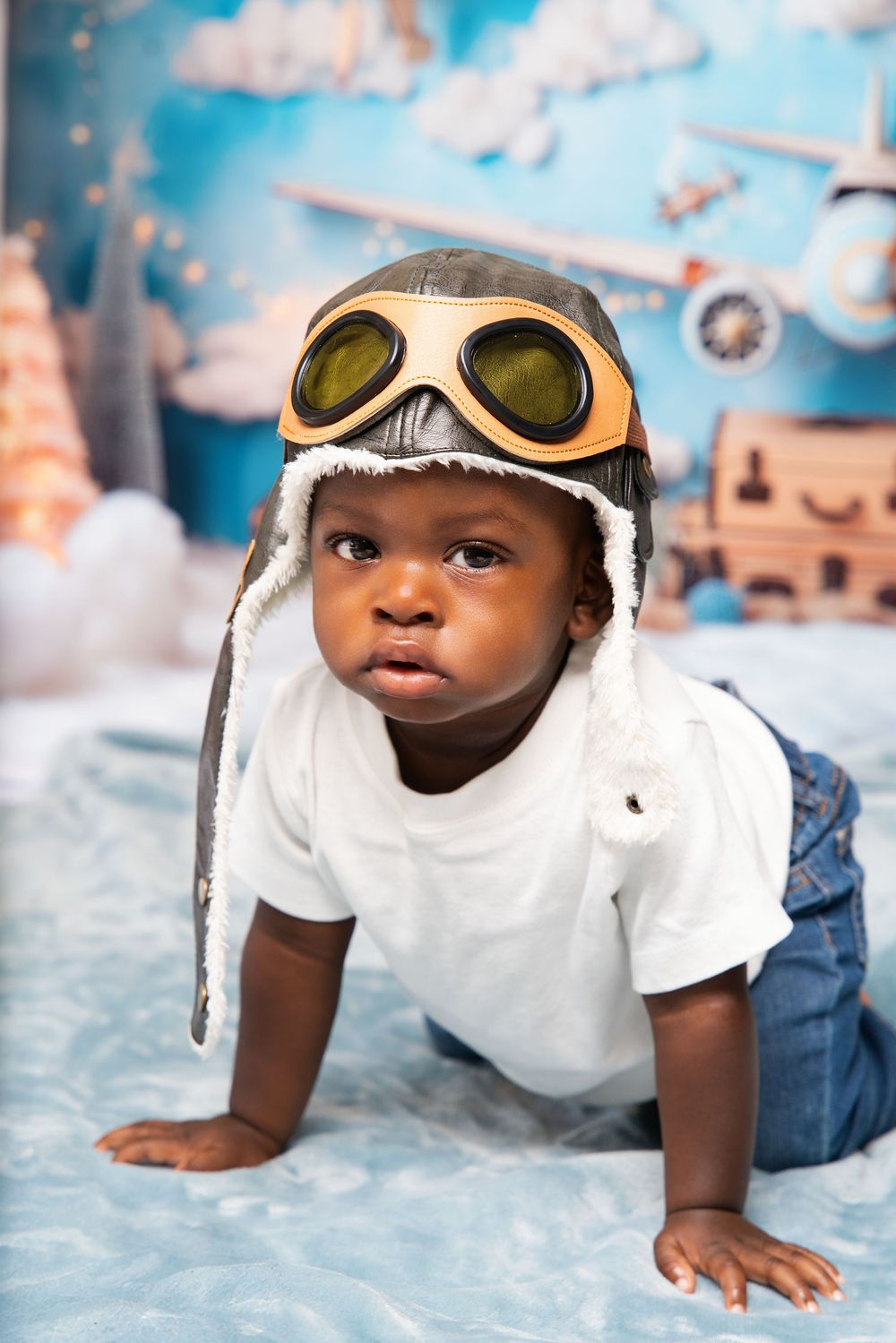 Adorable Black baby wearing vintage aviator hat with goggles, crawling on blue surface with airplane themed backdrop.