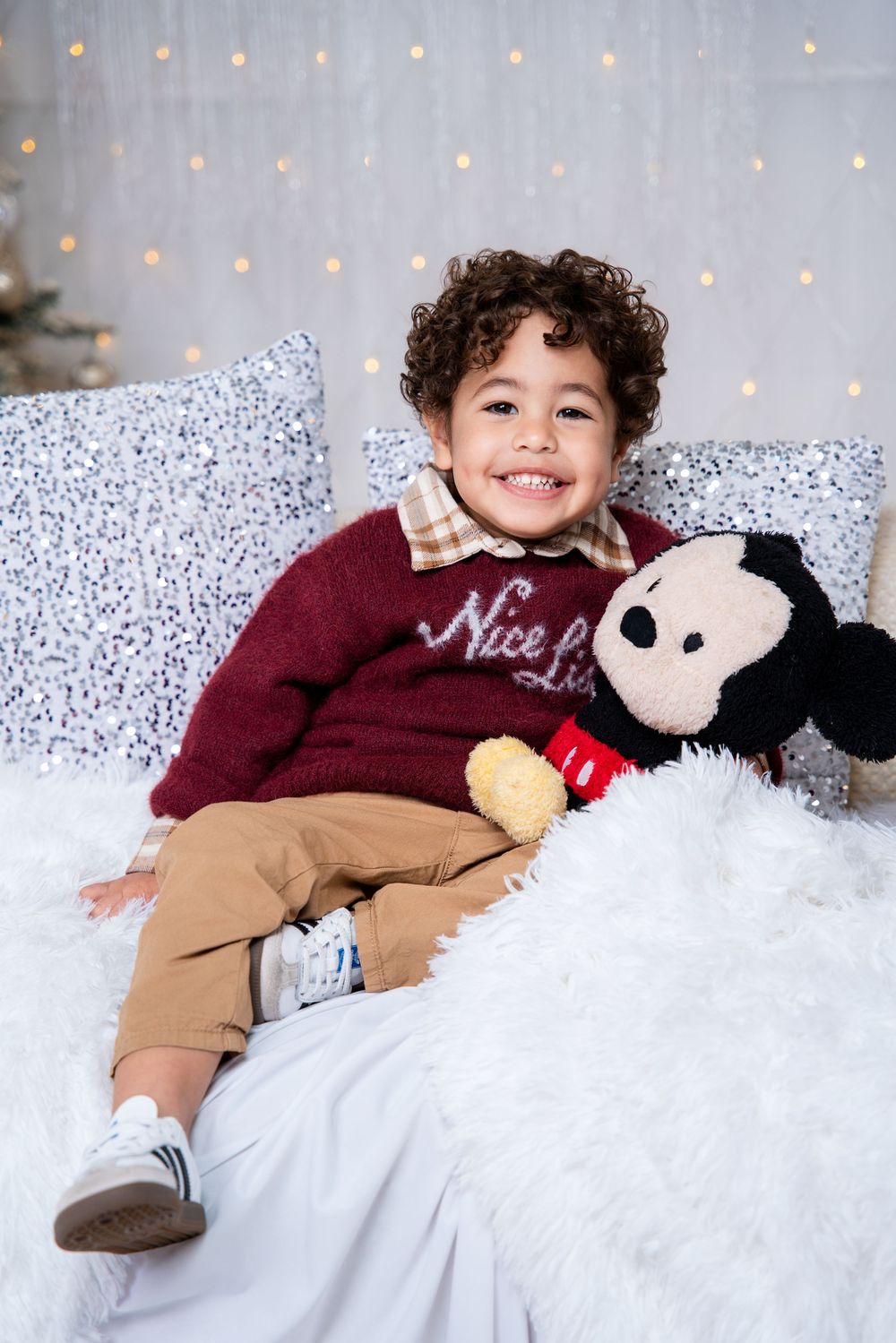 Child in burgundy sweater and khaki pants lies on white fluffy blanket with stuffed animal during holiday photo session.