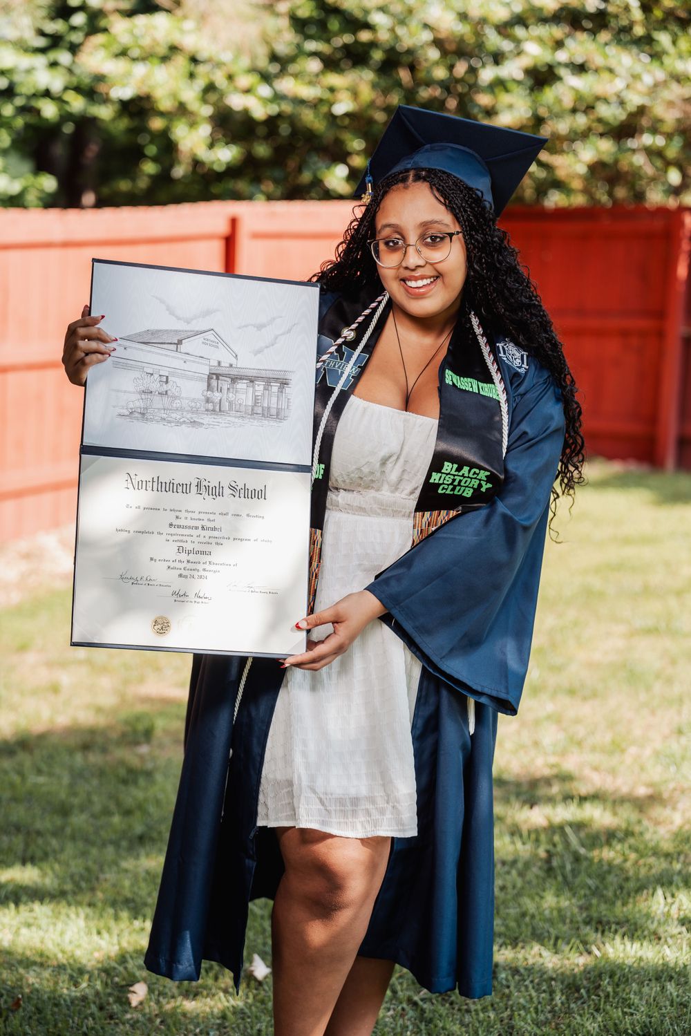 Graduate in blue cap and gown holds up diploma artwork and certificate during outdoor graduation ceremony.
