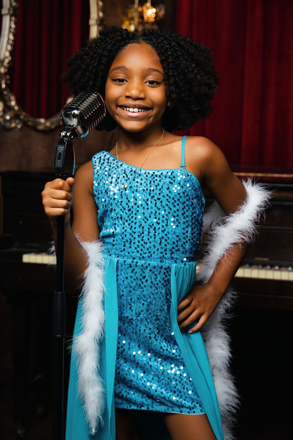 Young girl in blue sequin dress with feather boa sings into vintage microphone on stage, smiling confidently.