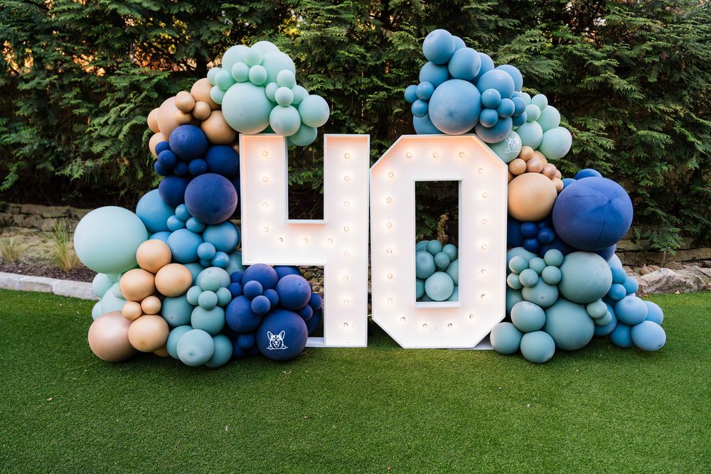 Marquee number 40 sign surrounded by blue, teal, and gold balloon garland on green grass for a 40th birthday.