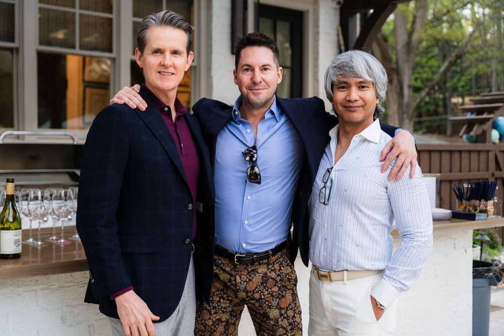 Three well-dressed men smiling and posing together at an outdoor social event with wine bottles visible in the background.