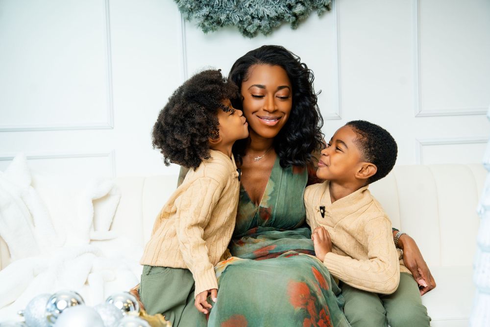 Sweet candid moment of children giving kisses to parent in front of white wall with wreath.