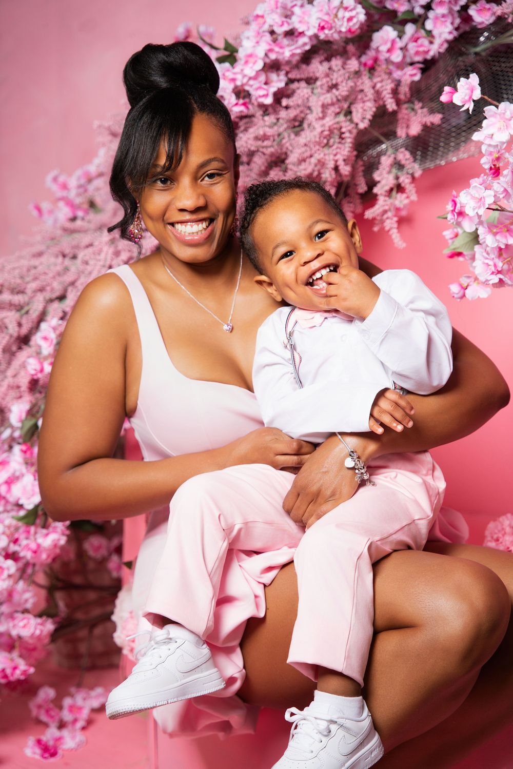 A smiling mother holds her happy baby, both dressed in pink and white, posed before a pink floral backdrop.