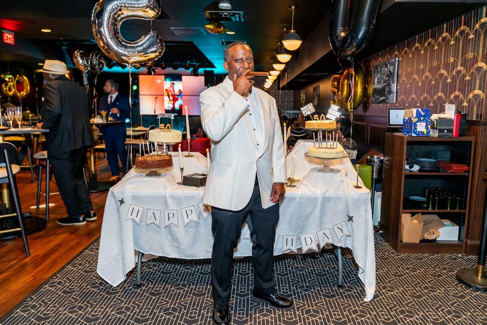 Man celebrating birthday party standing before Happy Birthday banner table with cake and balloons in restaurant.
