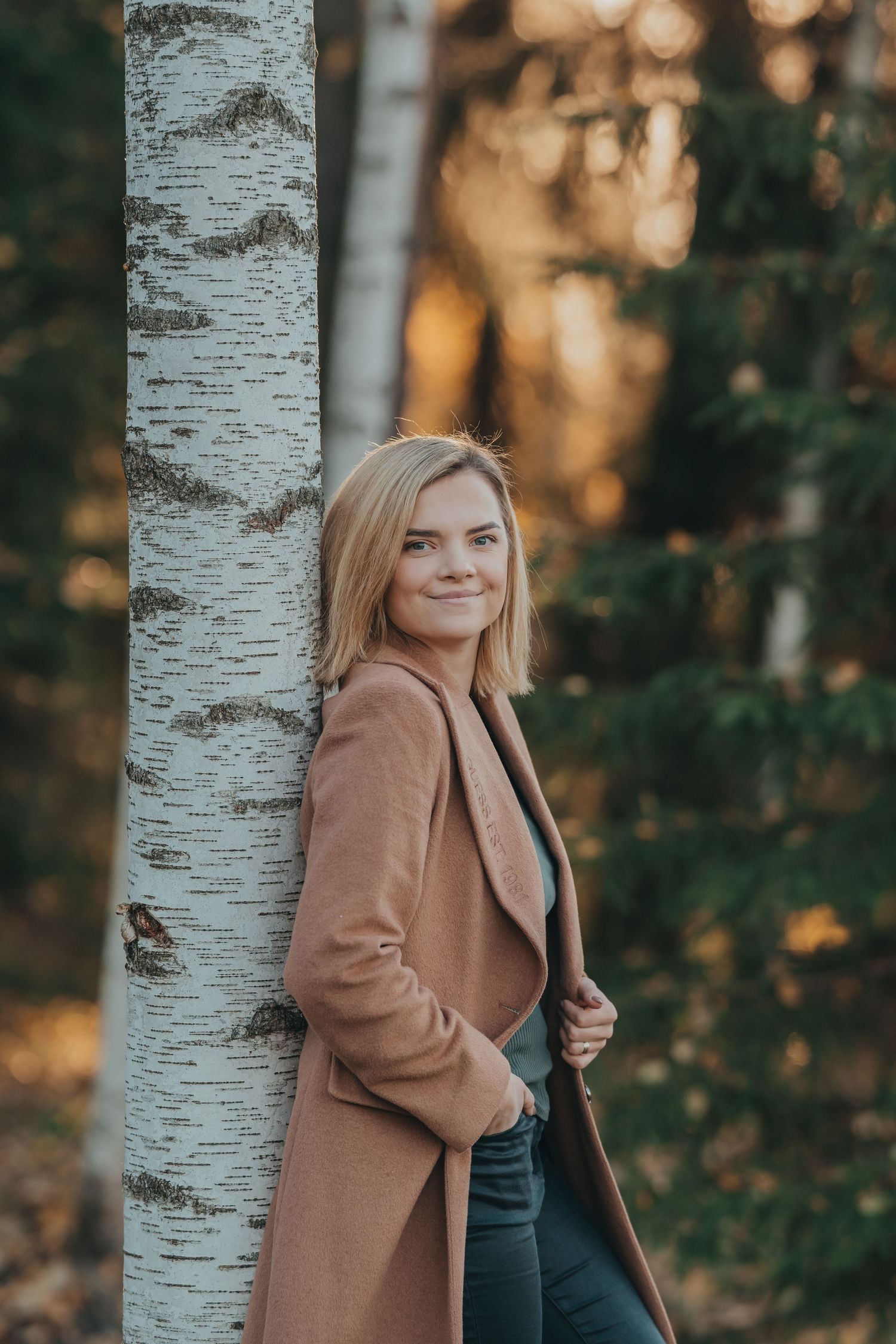 Portrait of someone in a camel coat leaning against a birch tree in autumn woodland.