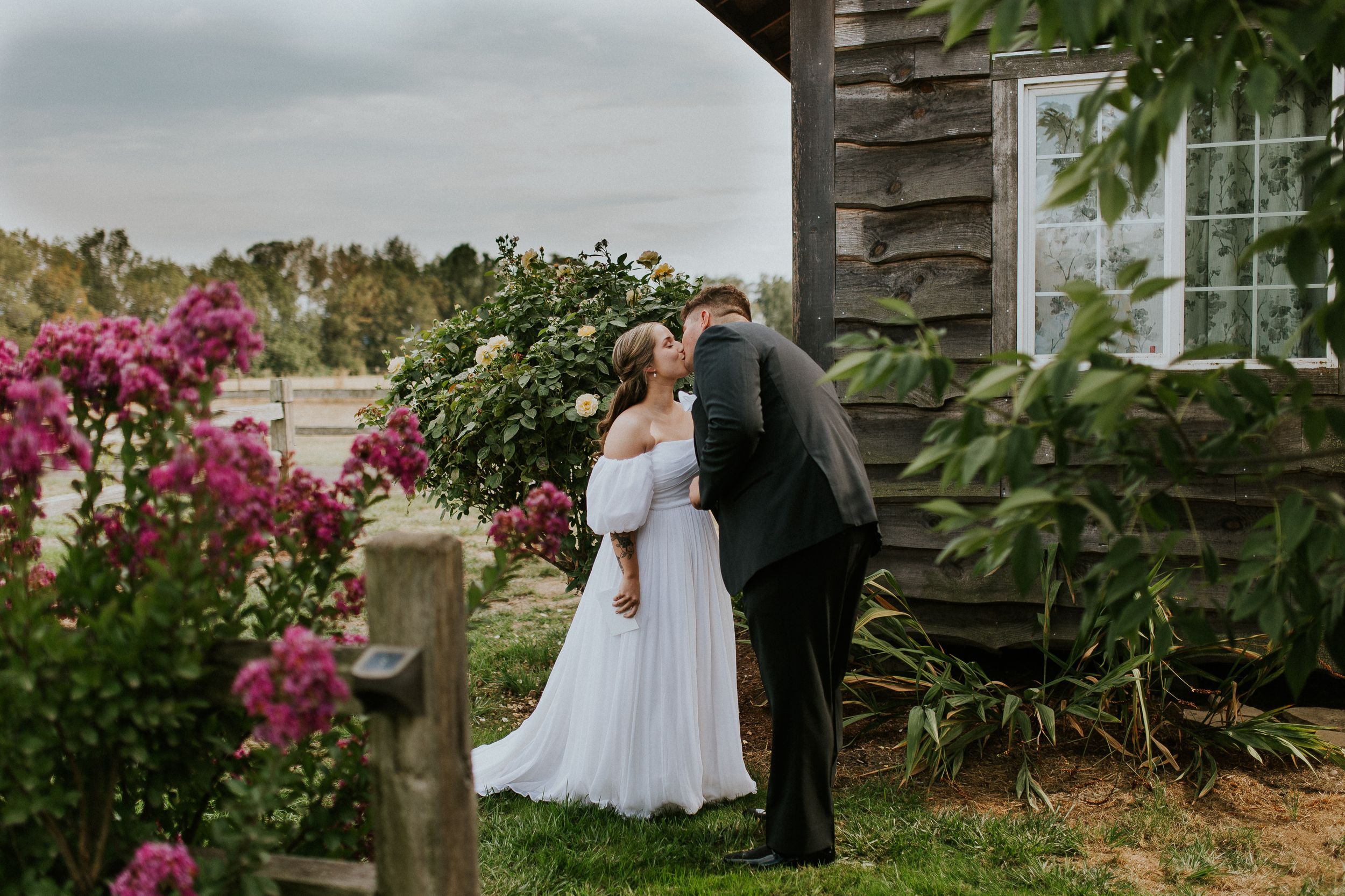 A couple in wedding attire embraces near a rustic building surrounded by colorful flowers and greenery. Creekside Farm wedding photography.