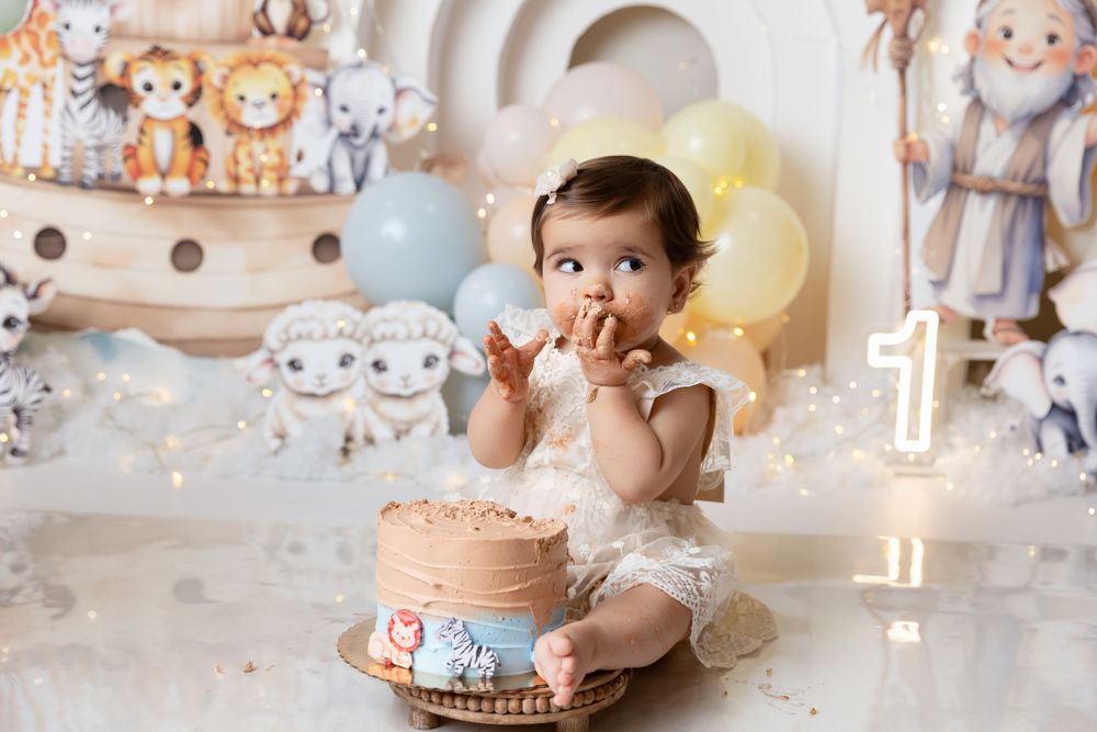 Baby in white outfit enjoys first birthday cake smash photo session with Noah's Ark themed decorations and balloons.
