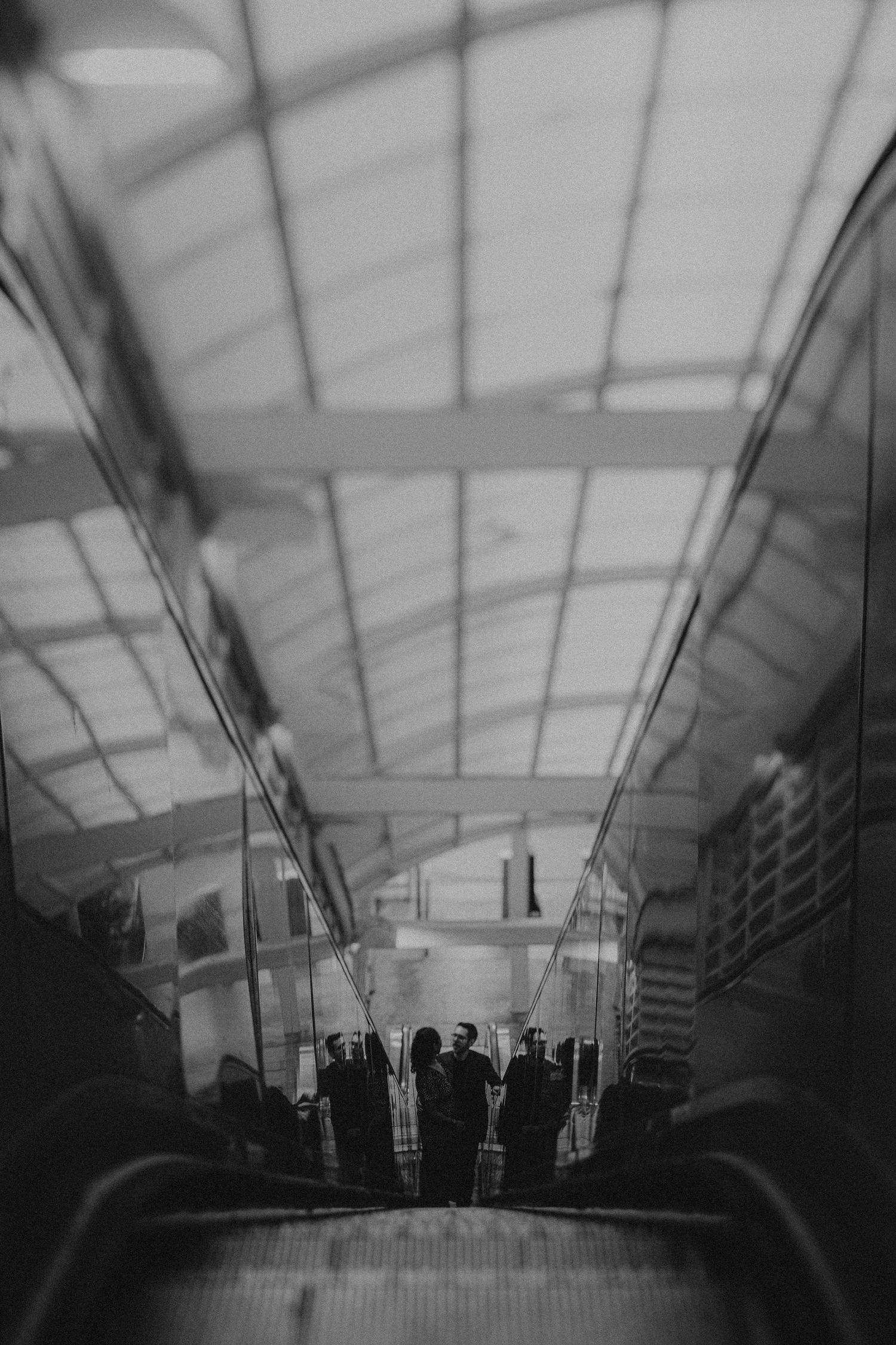 A black and white shot of a couple standing at the bottom of an escalator in a modern train station.
