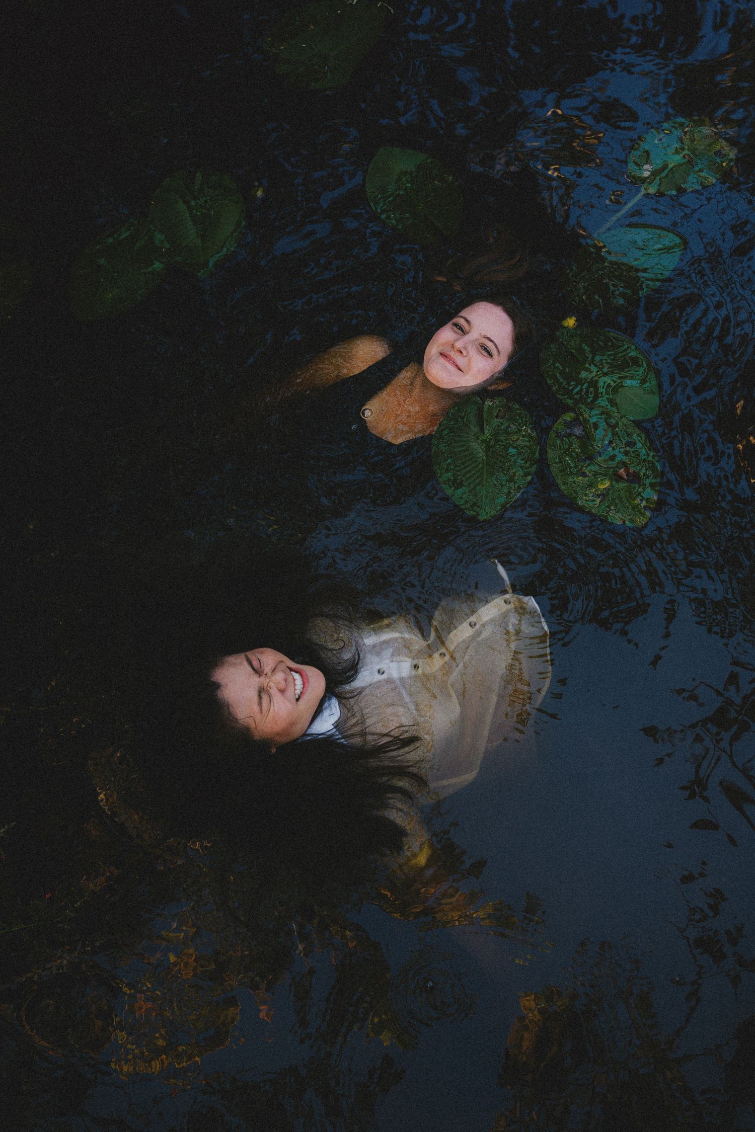 Artistic dark photo of a couple floating in water surrounded by lily pads and reflections at night.