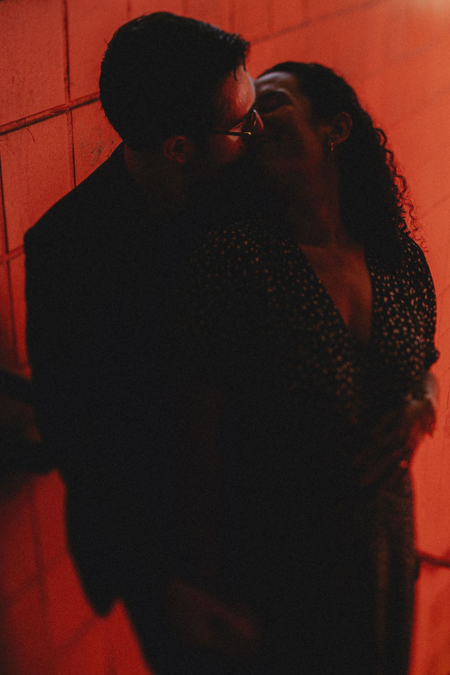 A romantic couple in an intimate moment against a red-lit wall, cast in moody shadows and warm lighting.