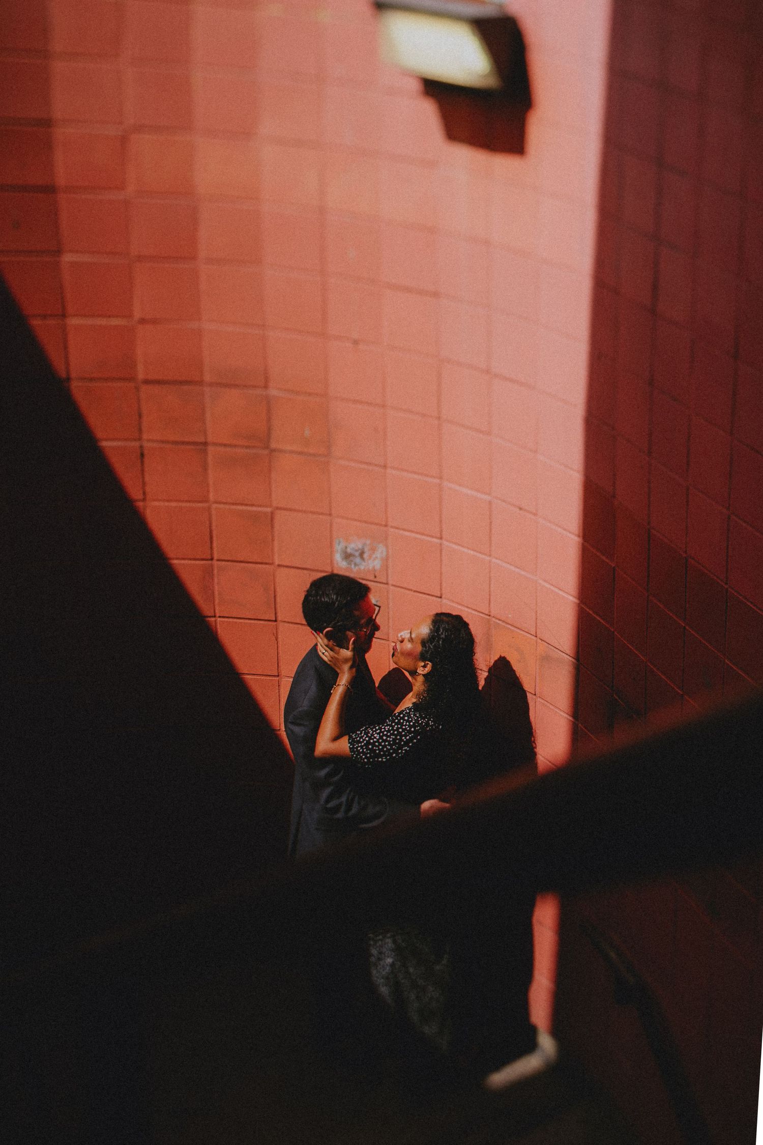 A figure emerges in shadows against a pink-lit brick wall creating a dramatic contrasting silhouette.