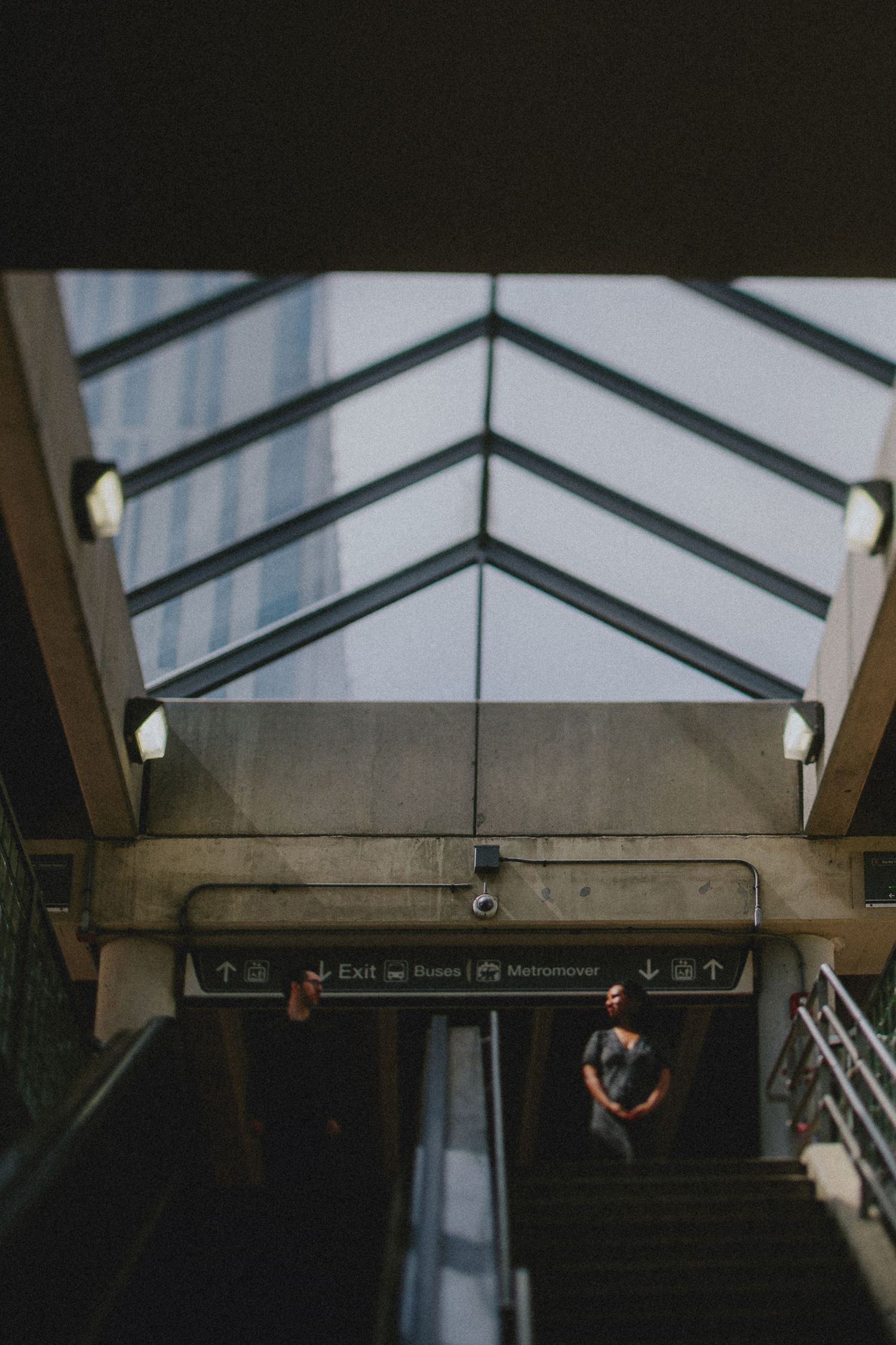 Dark moody subway station interior with geometric skylight pattern and couple silhouette figure waiting on platform below.