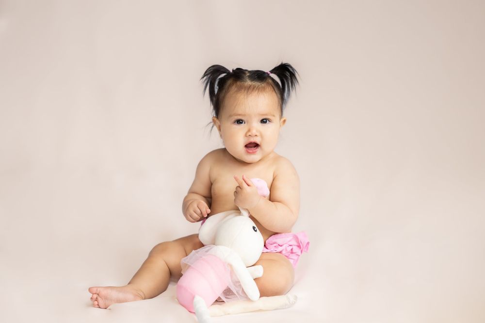 Baby sitting on white backdrop with pink stuffed animal wearing hair bows in a studio in Saratoga Springs.