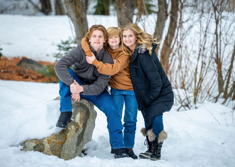 Three people huddle together for a winter portrait on snowy ground near trees and rocks.