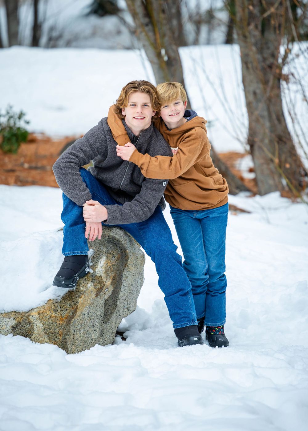 Two people smiling and posing together on a snowy winter day, with one sitting on a large rock and the other standing.