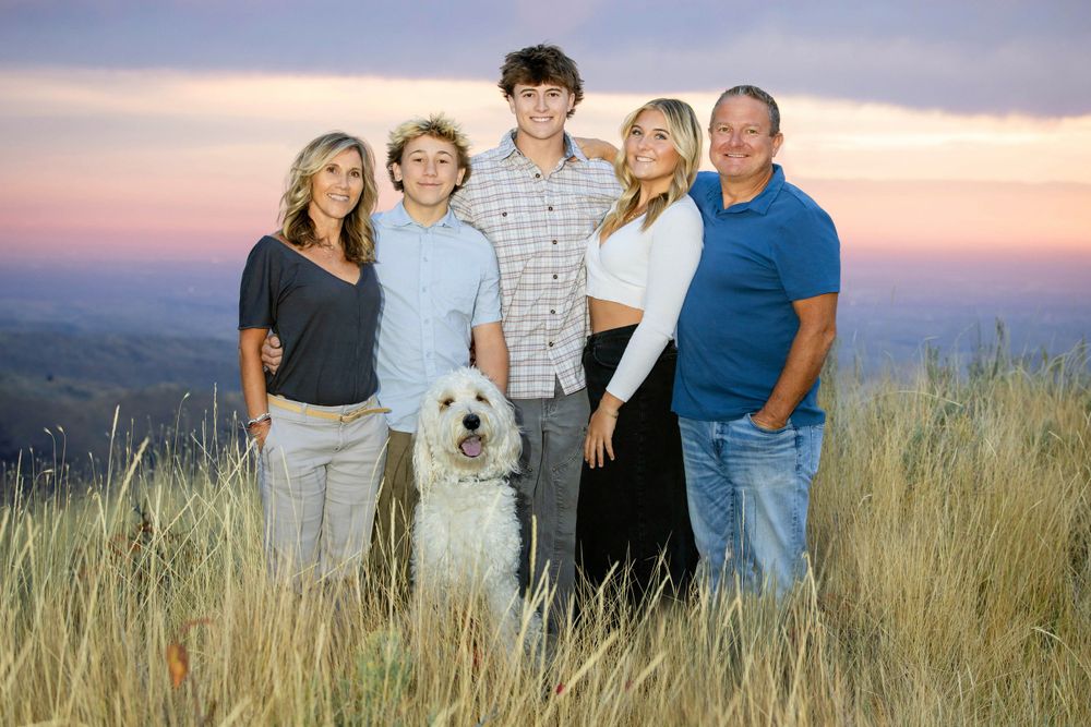 A family in casual attire stands together with their white dog against a mountain sunset backdrop in tall grass.