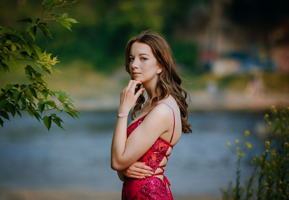 A young woman with long brown hair stands in a lush, green environment, wearing a red dress and looking thoughtfully to the side.