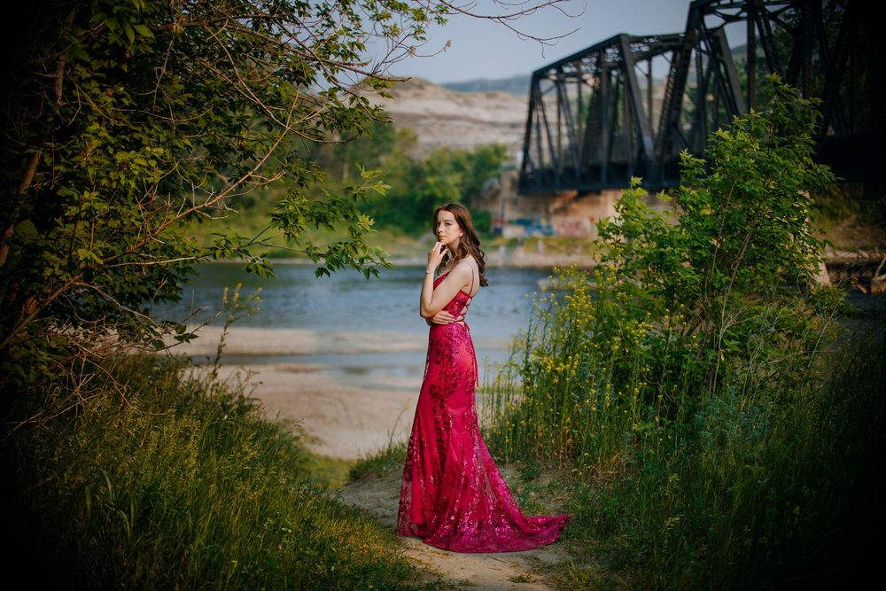 A woman in a long, red dress stands in a grassy field near a river and bridge.