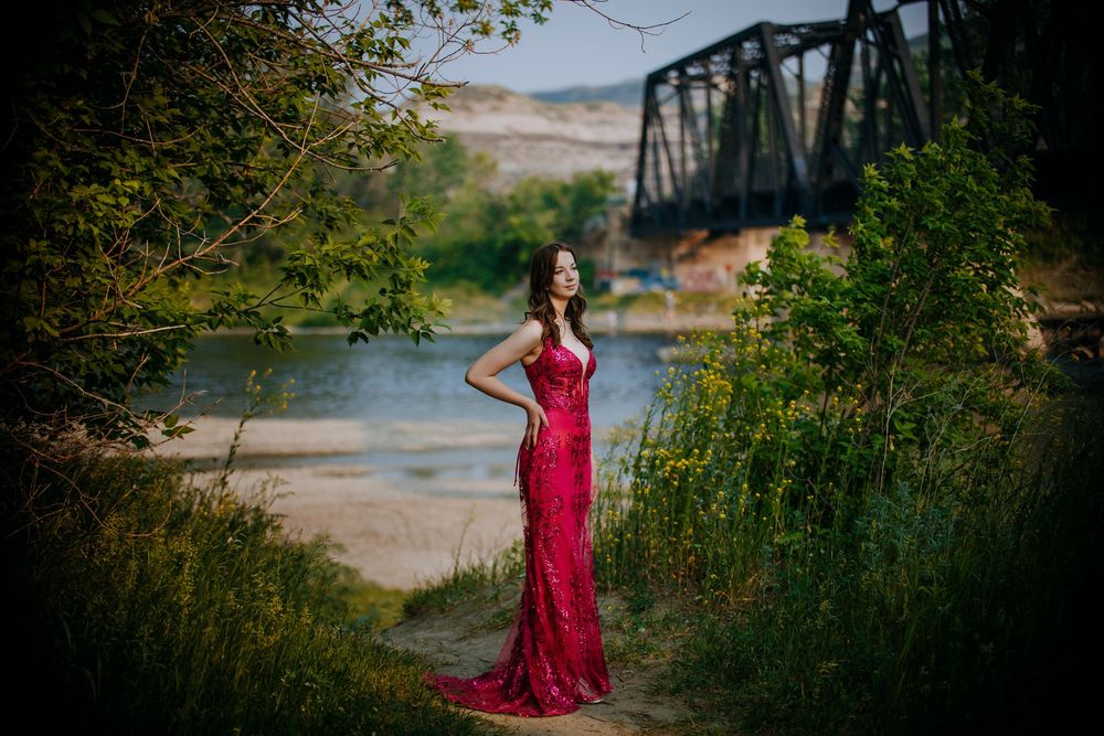 A woman in a long, red dress stands on a path near a river, with a bridge in the background.