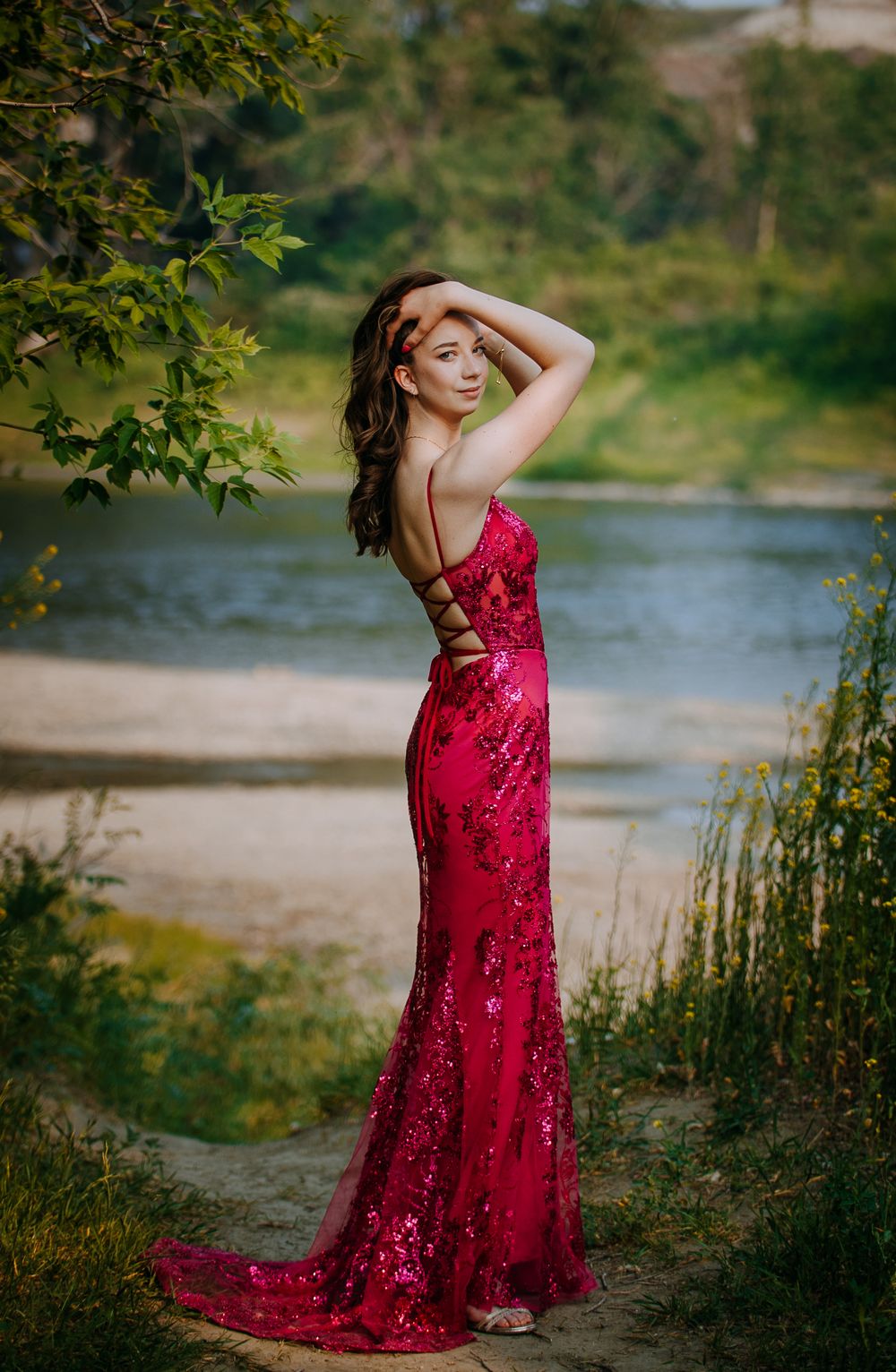 A woman in a long, red sequined dress poses gracefully in a scenic outdoor setting.