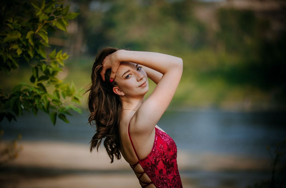 A woman in a red dress with a plunging neckline stands in a natural setting near a body of water.