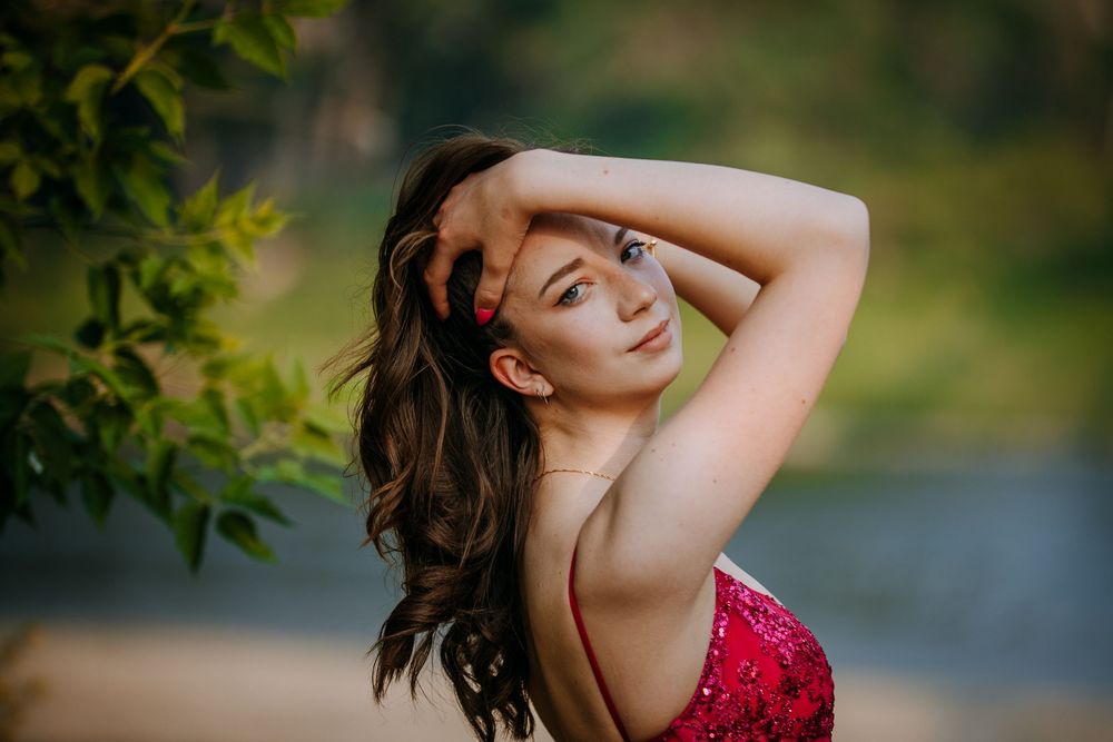 A woman with long brown hair wearing a red polka dot dress stands outdoors in a natural setting.
