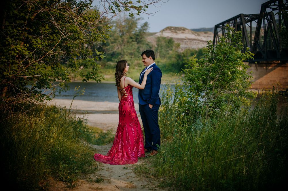 A couple in formal attire stand together in a grassy field near a river and bridge.