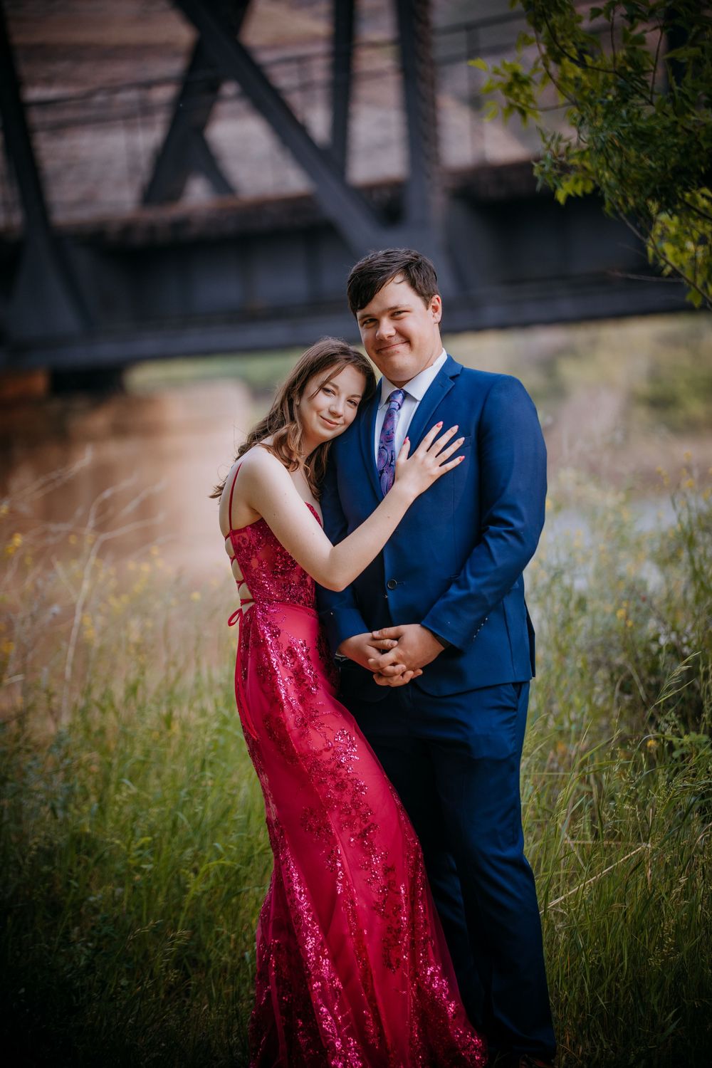 A couple in formal attire embracing each other in a scenic outdoor setting with a bridge in the background.