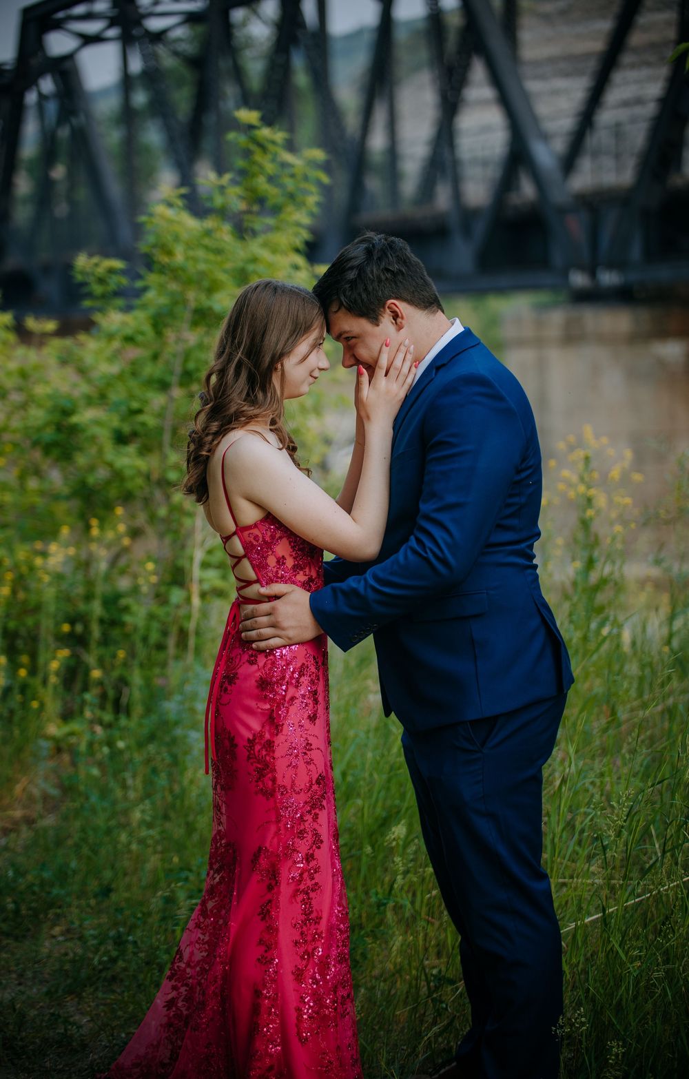 A couple in formal attire embraces tenderly in a scenic outdoor setting with a bridge in the background.
