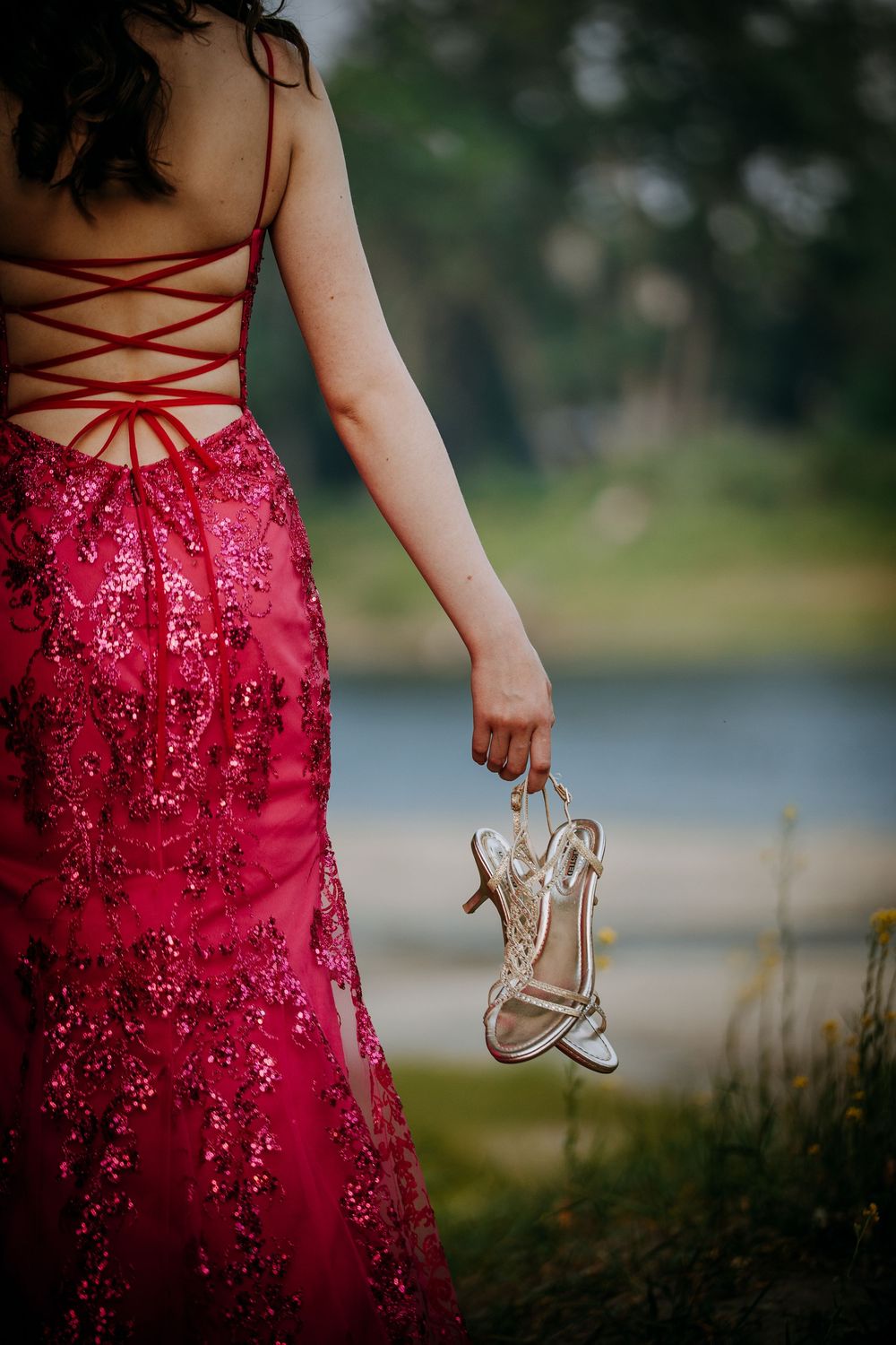 A woman in a red sequined dress holds a pair of ballet slippers in a natural outdoor setting.