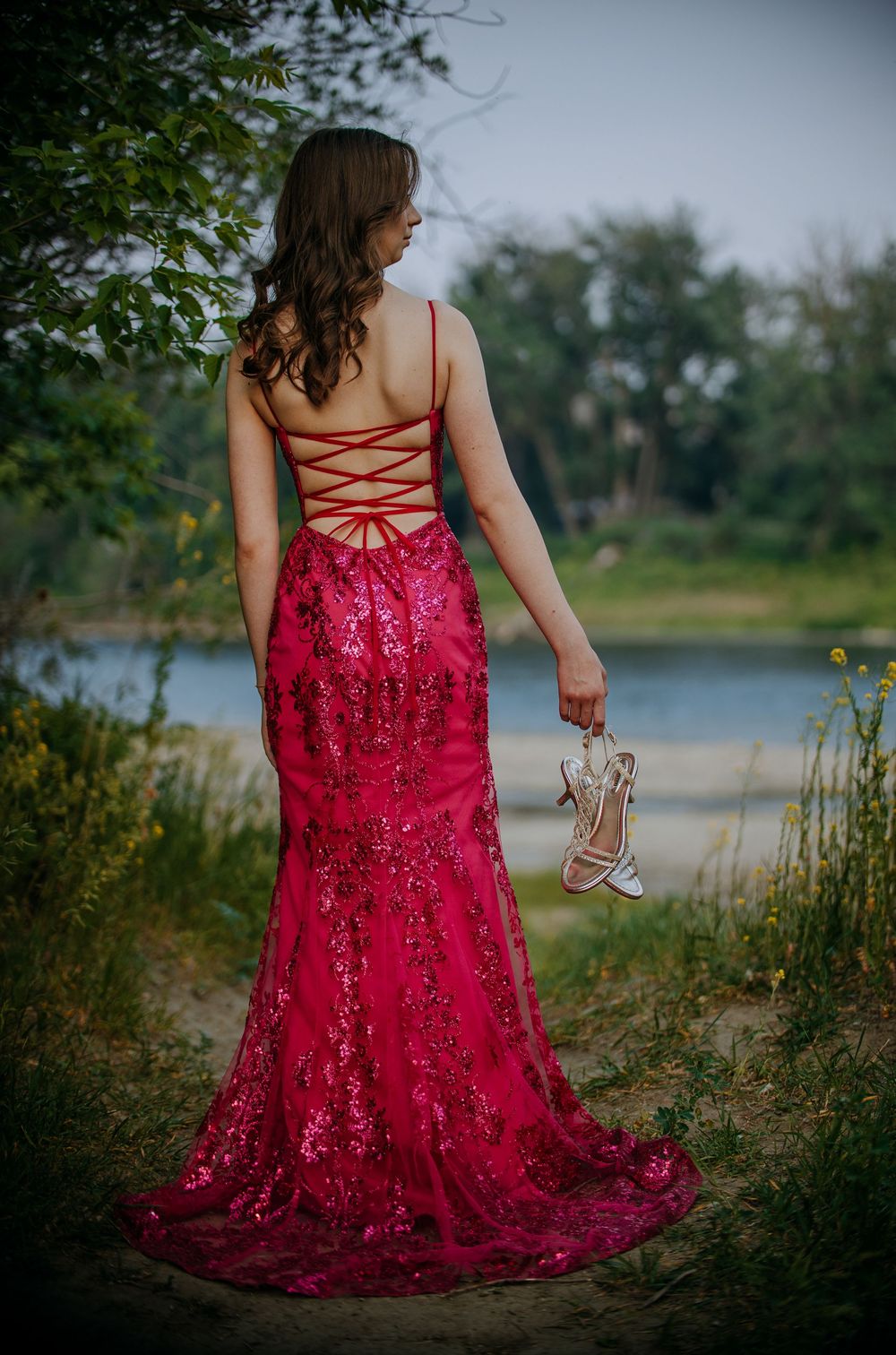 A woman in a long, red lace dress stands in a field near a body of water, holding a small bouquet of flowers.