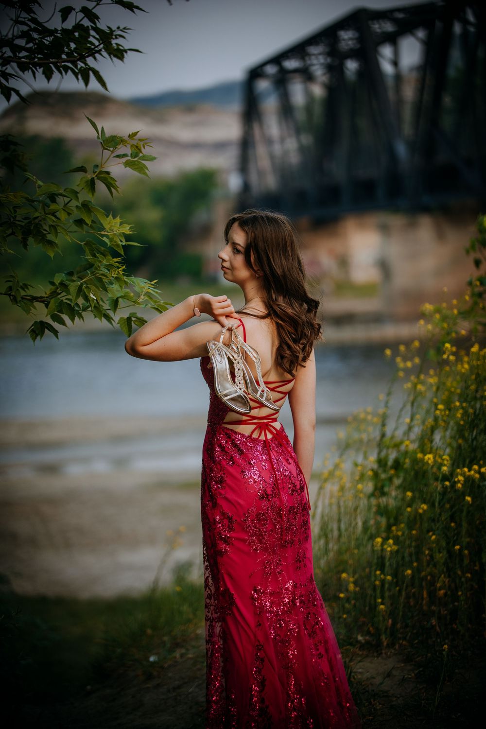 A woman with long brown hair in a red sequined dress stands in a scenic outdoor location with a bridge in the background.