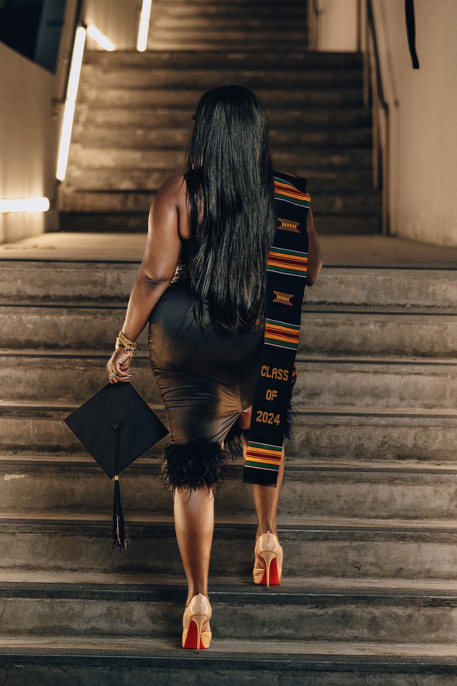 Graduate in black dress with Class of 2024 kente stole and cap walks up stone stairs.