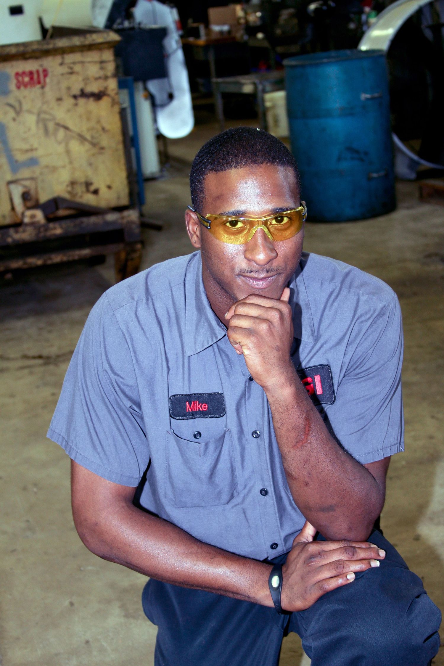 A worker in a grey uniform shirt and safety glasses poses thoughtfully in an industrial setting.