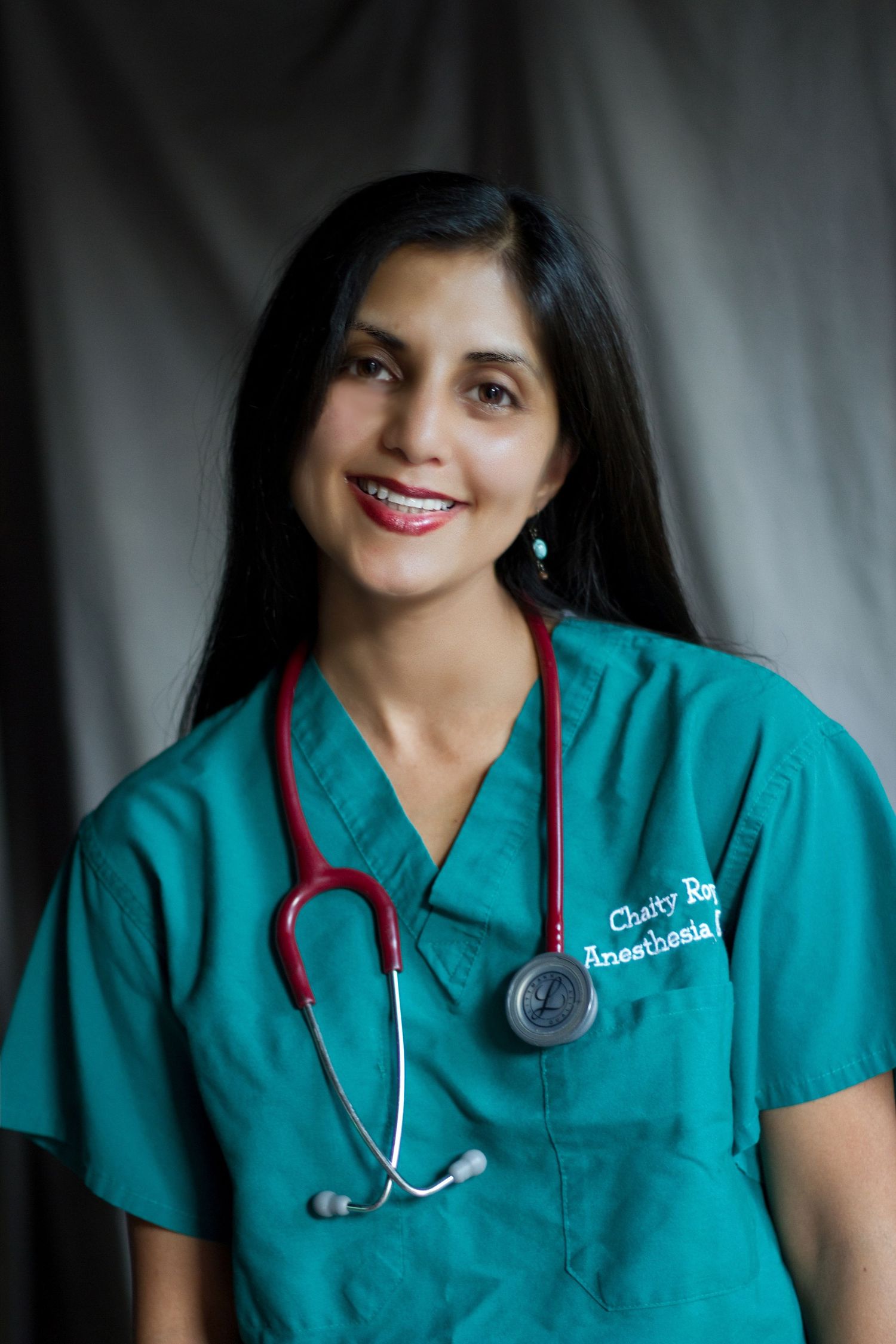 Healthcare professional wearing teal scrubs and stethoscope poses for a portrait against a grey backdrop.