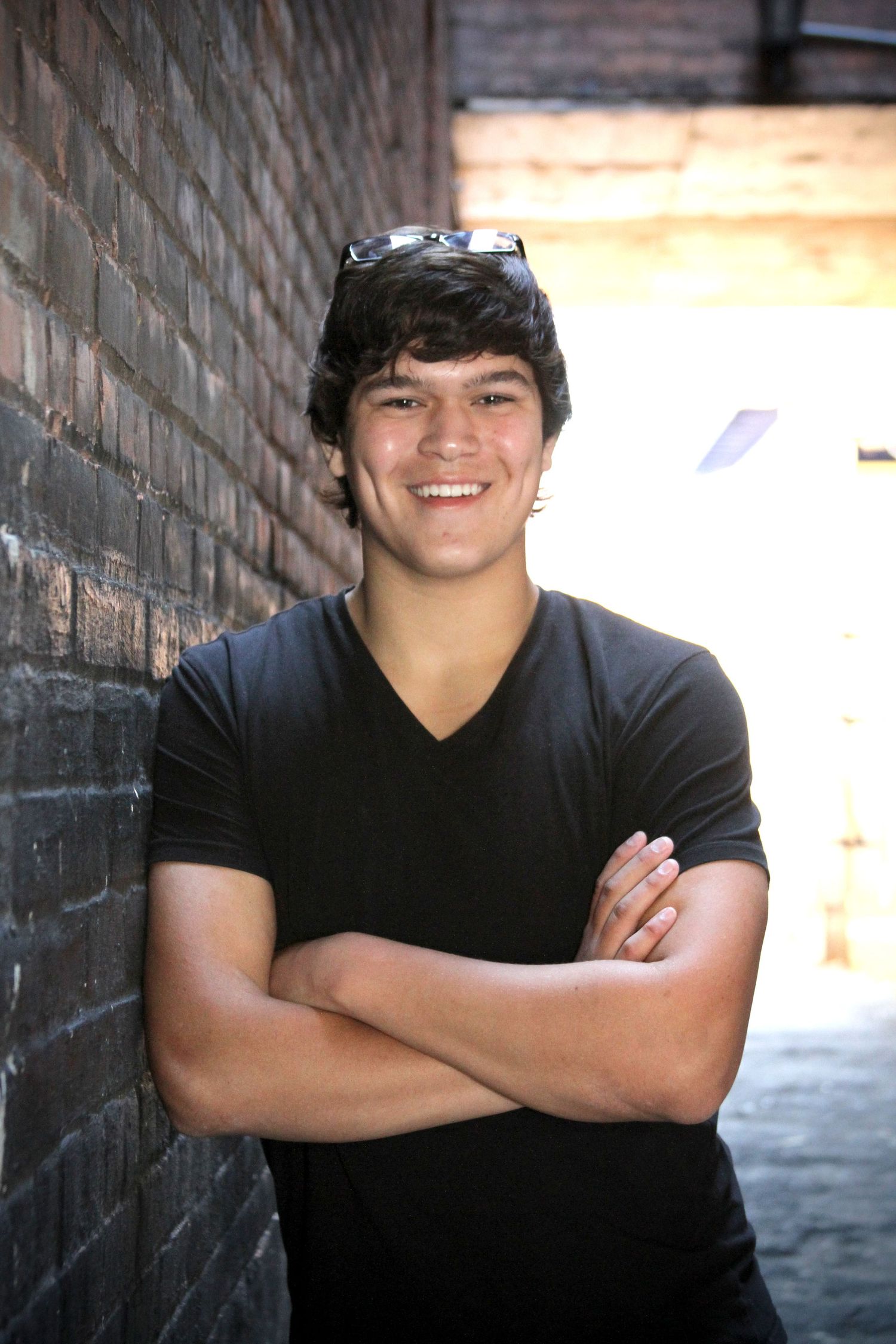 Young person in black v-neck t-shirt smiling against brick wall with sunglasses on head.