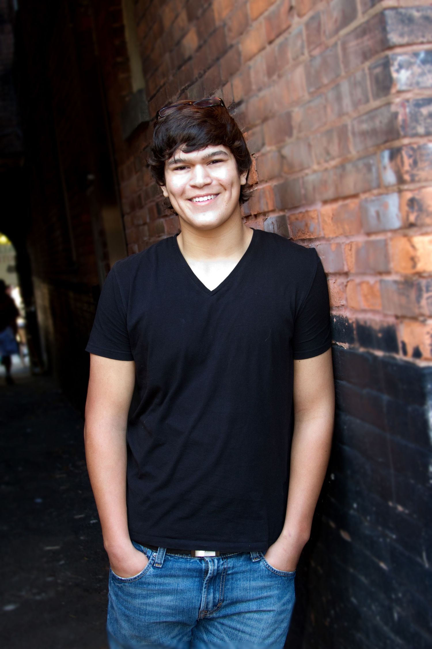 Casual portrait of person in black v-neck shirt standing against brick wall background.