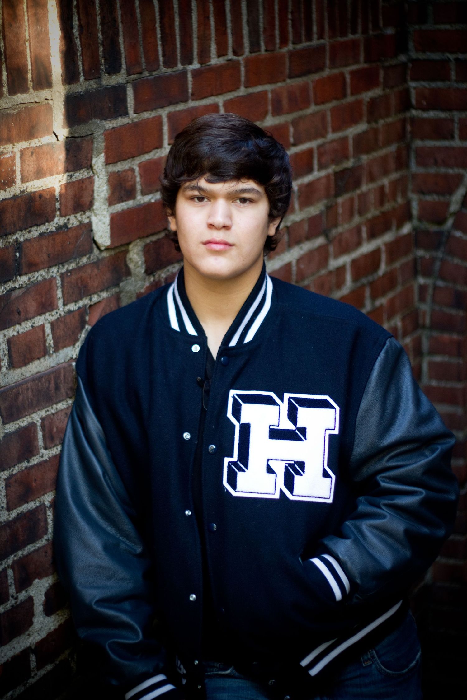 Person wearing varsity letterman jacket with 'H' logo poses against brick wall.