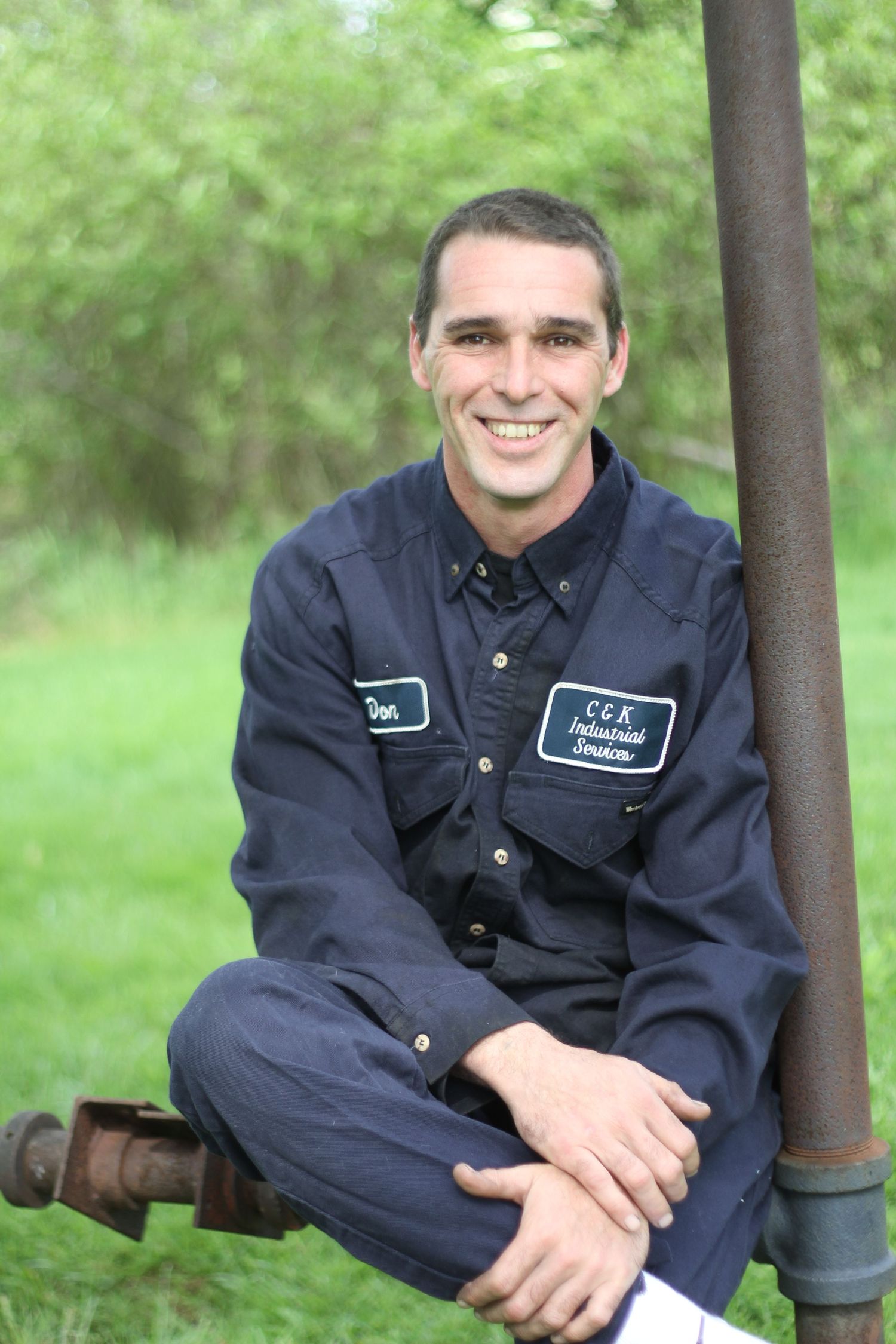 Maintenance worker in navy uniform sits casually on industrial equipment outdoors.