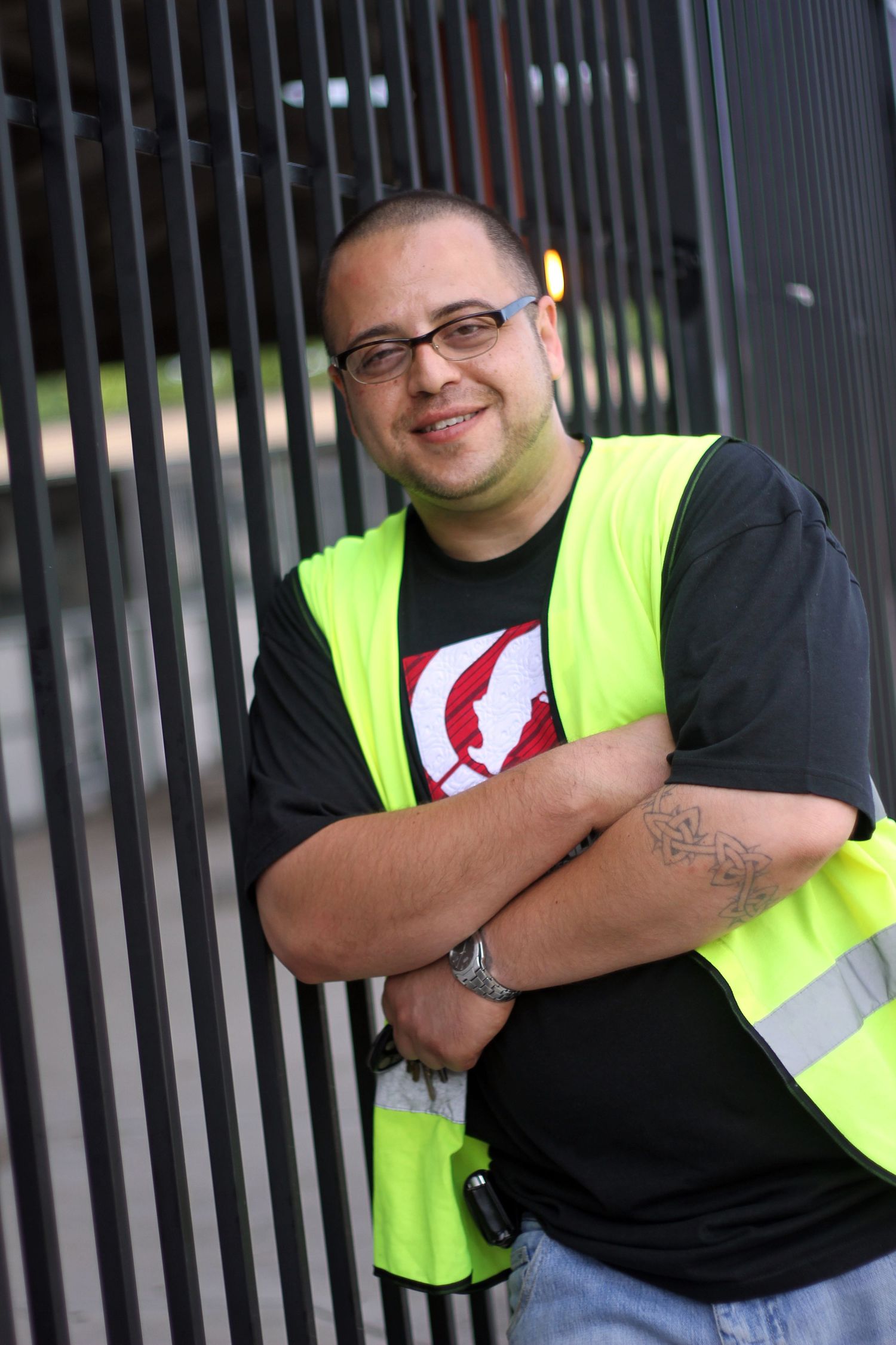 Safety worker in high visibility vest and glasses smiles while standing near metal gate.