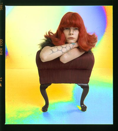 Artistic portrait with vibrant rainbow background featuring person with bright red hair behind furniture.