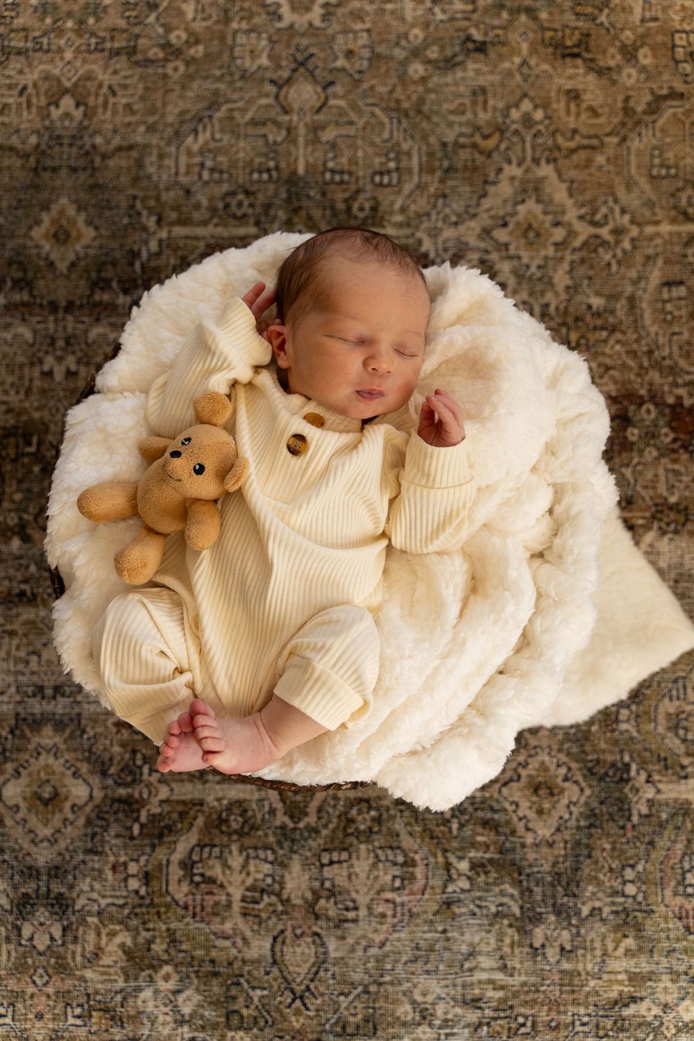 Sleeping newborn in cream knit outfit lies on soft white blanket with small teddy bear on patterned carpet background.