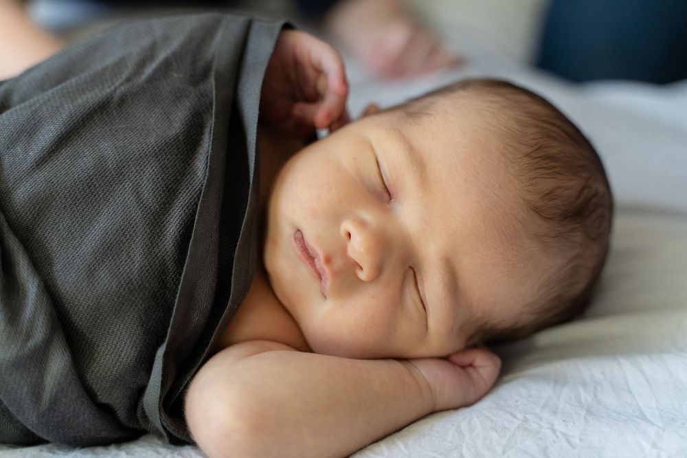 A peaceful sleeping baby wrapped in a gray blanket rests on white bedding in natural light.