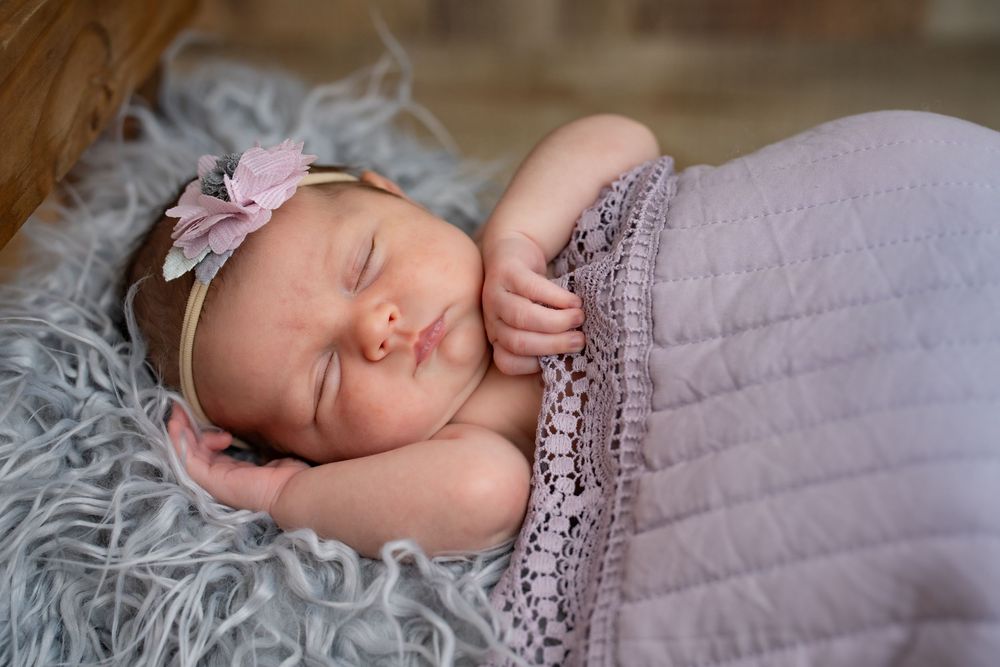 A sleeping newborn swaddled in soft lilac blanket with lace trim rests on fluffy gray fur backdrop.