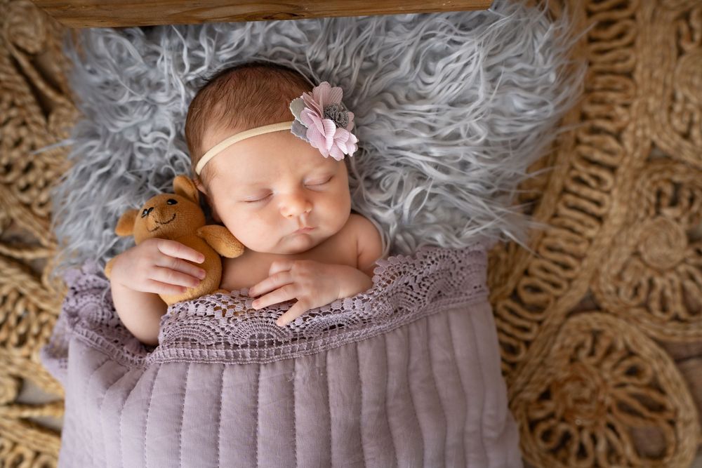Sleeping newborn baby wrapped in purple knit blanket on fluffy gray rug with pink floral headband.