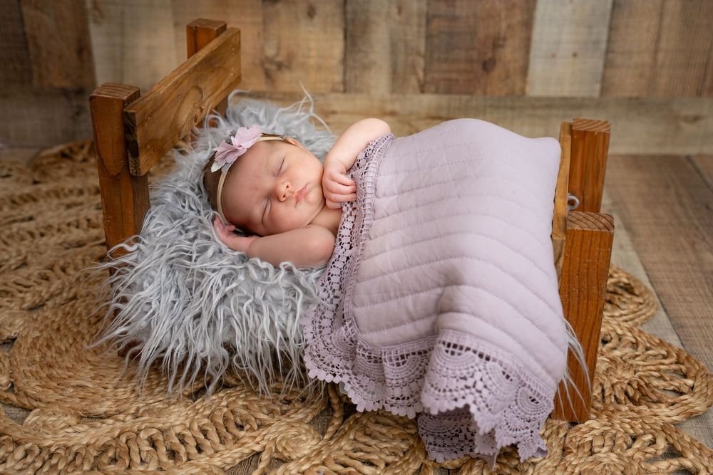 A sleeping baby rests on a soft gray fur cushion in a miniature wooden bed with a lilac knit blanket.